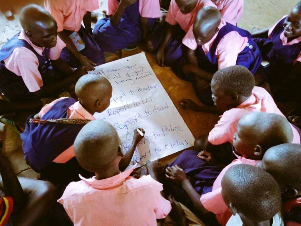 A circle of schoolchildren in pink uniforms filling in a flipchart on how to report abuse