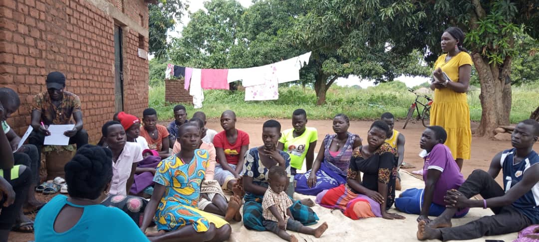 A community outreach session with a facilitator addressing a group of women and youth seated outdoors
