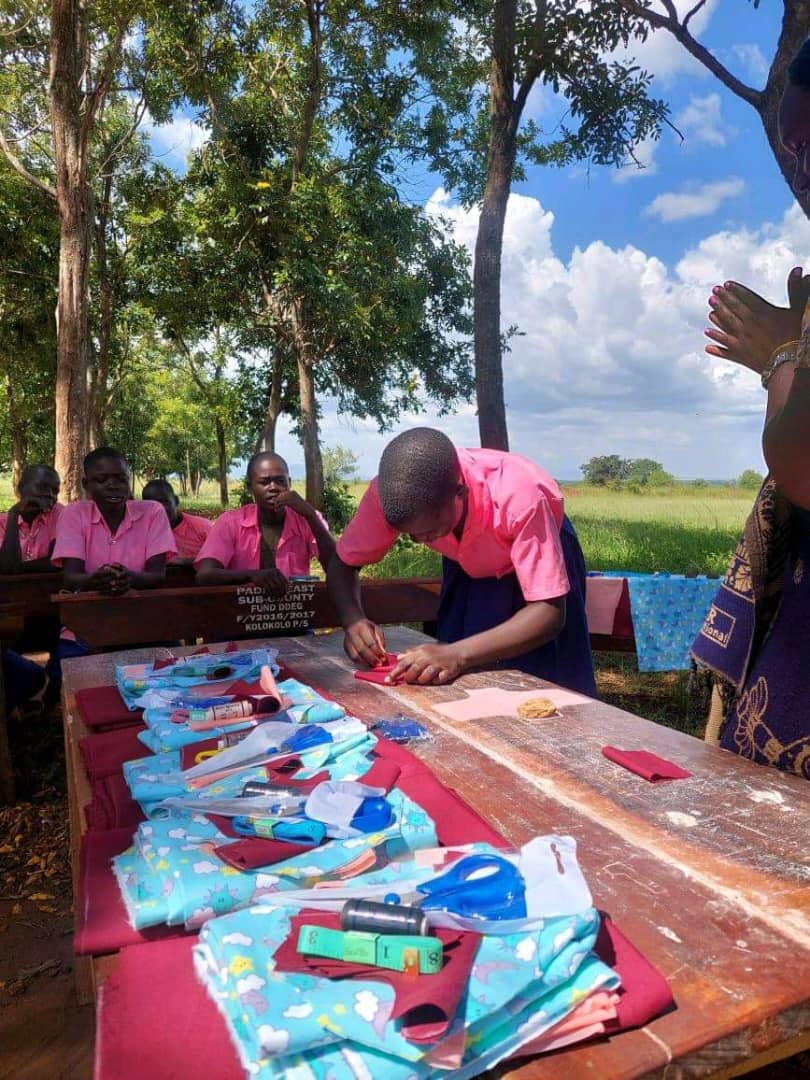 A schoolgirl in a pink uniform sewing a reusable sanitary pad at an outdoor menstrual hygiene training